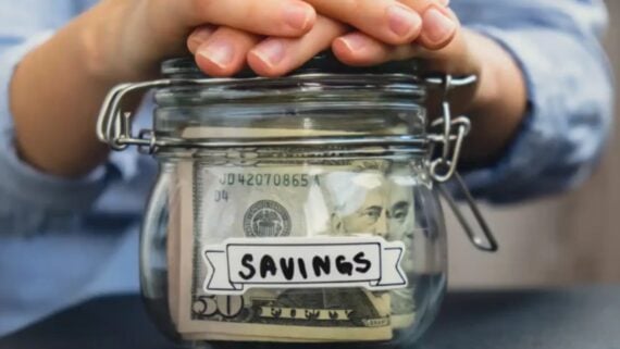 A person&rsquo;s hands rest on the lid of a glass jar labeled &ldquo;SAVINGS,&rdquo; which is filled with U.S. dollar bills.