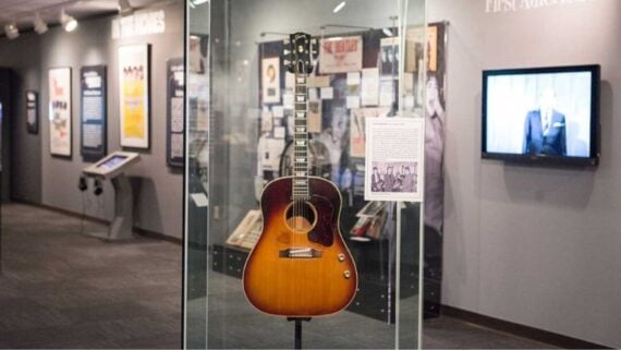 A sunburst acoustic guitar is displayed in a glass case at a museum exhibit, with music memorabilia and framed photos on the walls and a TV screen in the background.