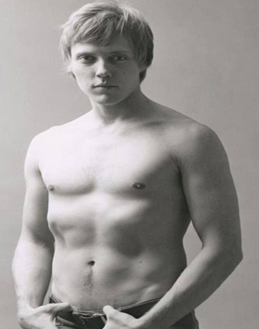 A young, light-skinned man stands shirtless against a plain background, looking directly at the camera with a neutral expression. He has short, light hair and is holding the waistband of his pants.