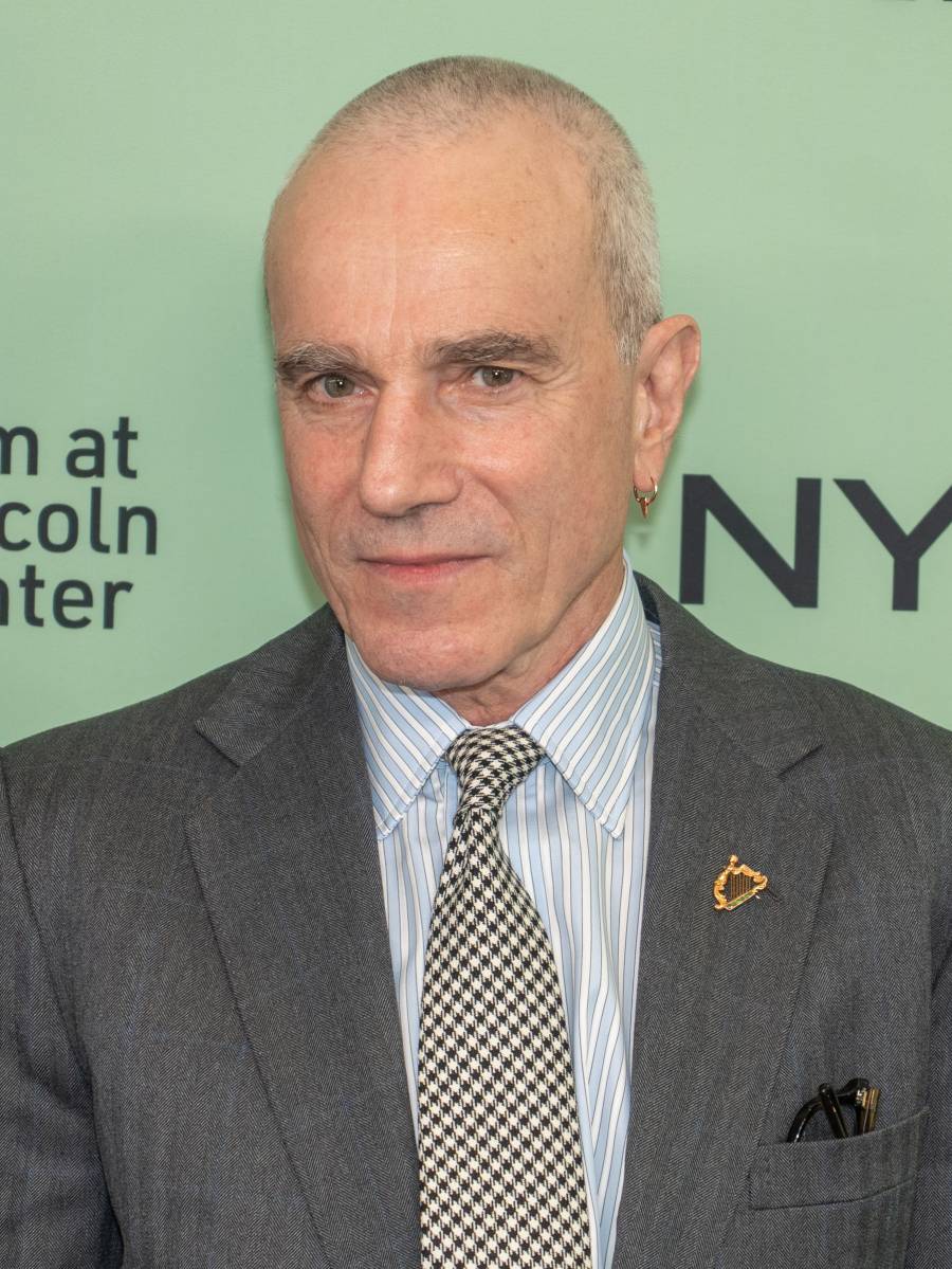 A man with closely cropped gray hair wearing a gray suit, striped shirt, and checkered tie, standing in front of a green background with partially visible text.