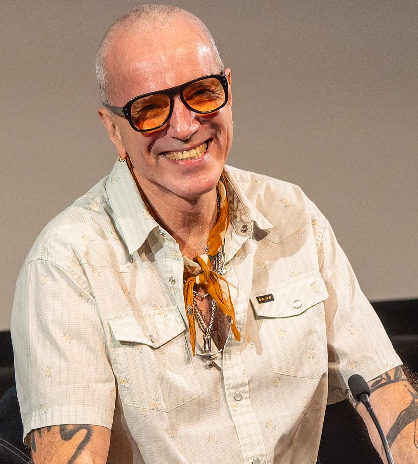 A smiling person with short hair and sunglasses, wearing a beige shirt with front pockets and an orange scarf around their neck, sits in front of a microphone.