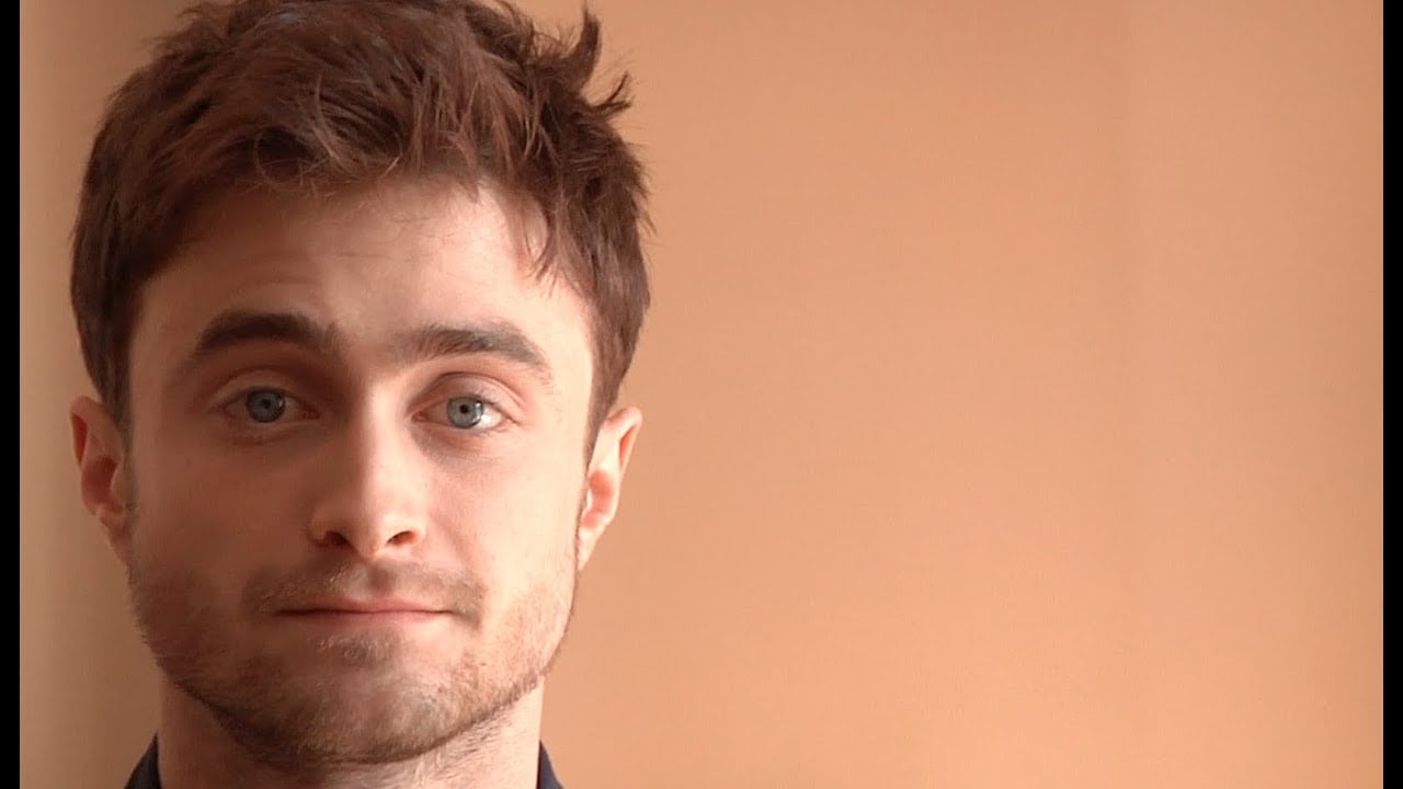 A young man with short brown hair and blue eyes looks into the camera with a slight smile, standing in front of a plain beige background. His face is partially lit from the left side of the image.