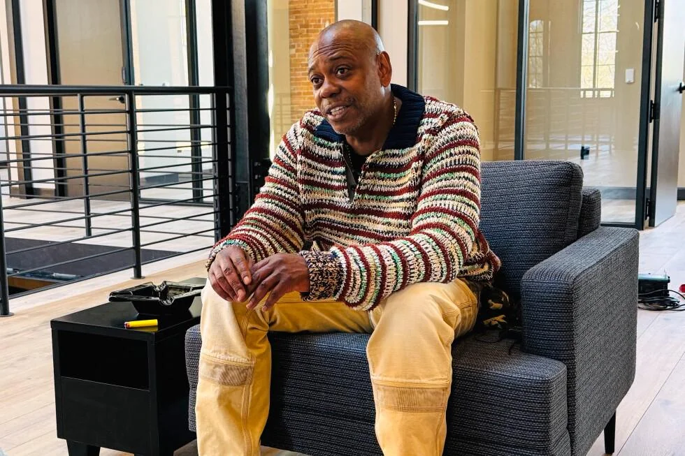 A man in a striped sweater and tan pants sits on a gray armchair in a modern, well-lit office space, smiling and leaning forward with his hands clasped.