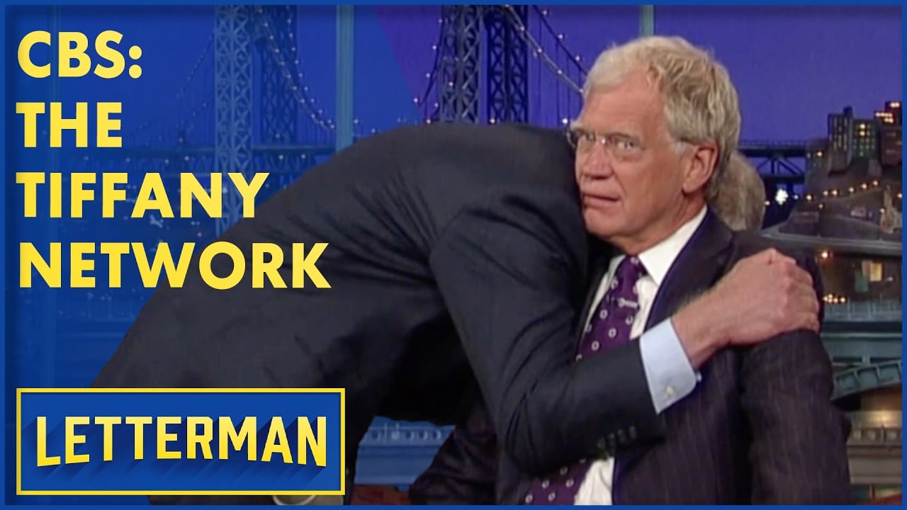 Two men sit on a late-night talk show set. One man hugs the other, whose expression looks surprised. Yellow text reads "CBS: THE TIFFANY NETWORK" and "LETTERMAN" appears in a blue and yellow box at the bottom.