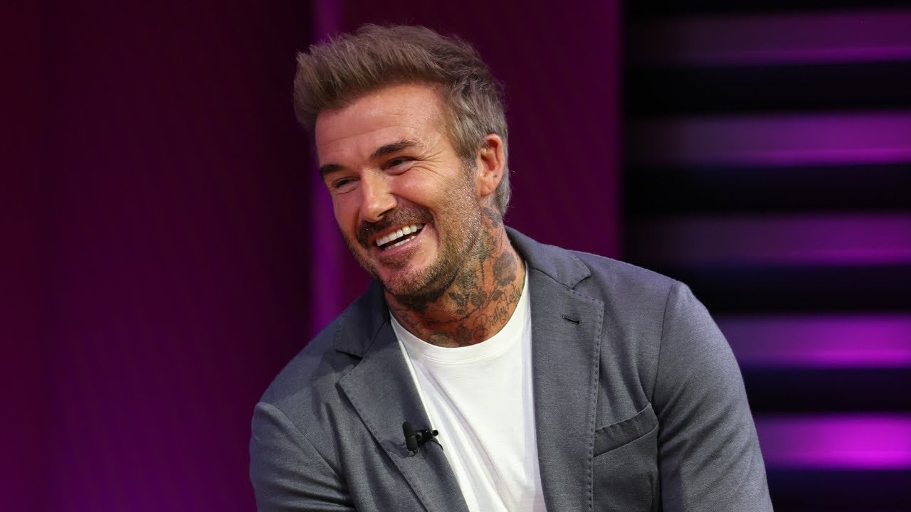 A man with light brown hair and tattoos on his neck is smiling while wearing a grey blazer over a white t-shirt, sitting against a purple background.