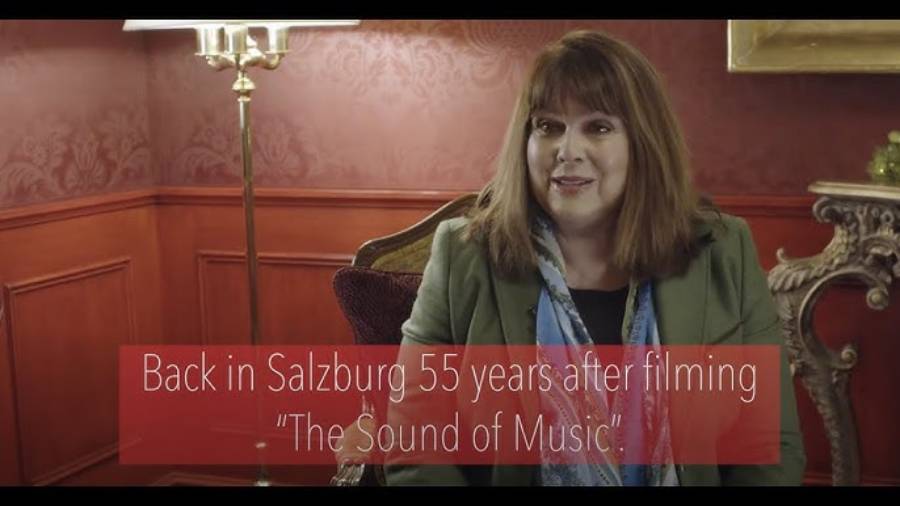A woman sits in an elegant room with red wallpaper and ornate furniture. Text on the image reads: "Back in Salzburg 55 years after filming 'The Sound of Music'.