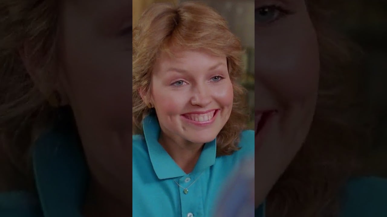 A woman with wavy blonde hair and a bright smile wears a turquoise collared shirt, looking slightly to the left. The background is softly blurred.