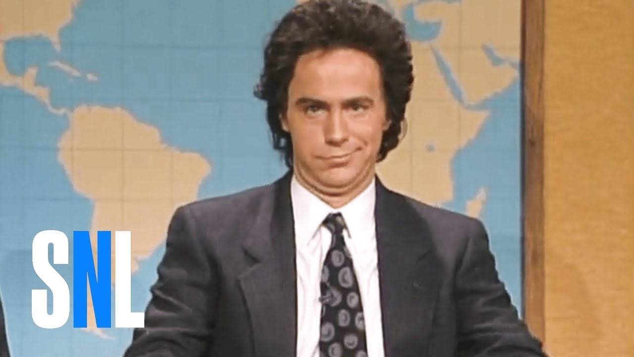 A person in a suit and tie sits at a news desk in front of a world map backdrop, with an amused facial expression. The "SNL" logo appears in the lower left corner.