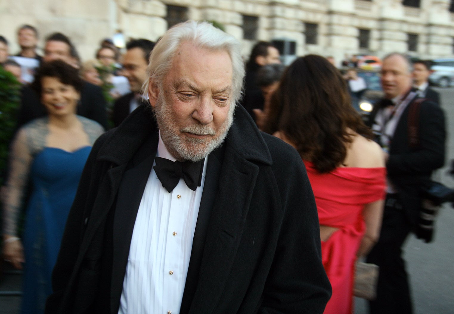 An older man with white hair and a beard, dressed in a tuxedo and black overcoat, walks outside at a formal event, surrounded by people in evening attire. The background shows a stone building and blurred attendees.