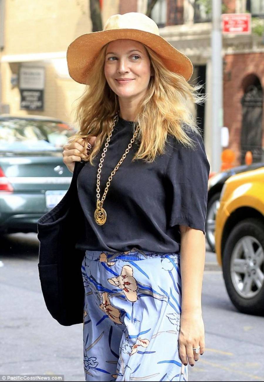 A woman with long blonde hair wearing a wide-brimmed straw hat, a black top, a gold chain necklace, and a floral skirt walks outdoors, smiling. Cars and buildings are visible in the background.