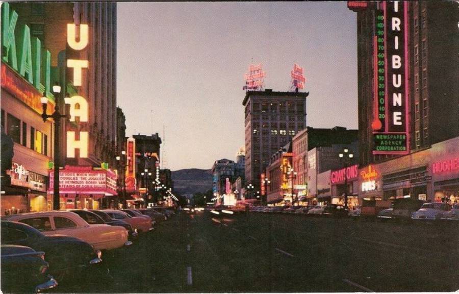 A vintage photo of a city street at dusk with neon signs for businesses like UTAH, KALL, and TRIBUNE, and classic cars parked along both sides of the road. Distant mountains are visible in the background.