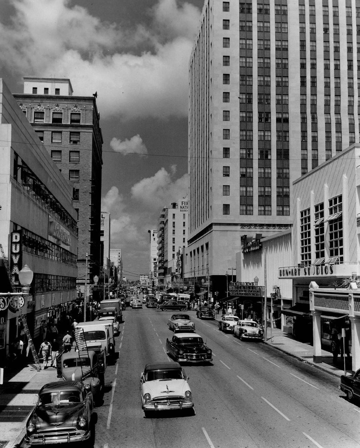 Black and white photo of a busy city street in the 1950s, with classic cars, people walking on sidewalks, and tall buildings lining both sides of the road under a partly cloudy sky.