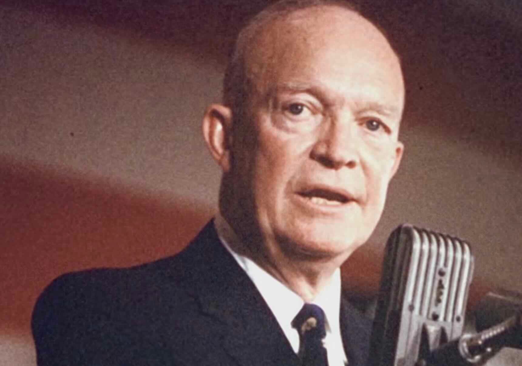 A man in a suit speaks into a vintage microphone, with part of an American flag visible in the background. He has a serious expression and is lit from the front.