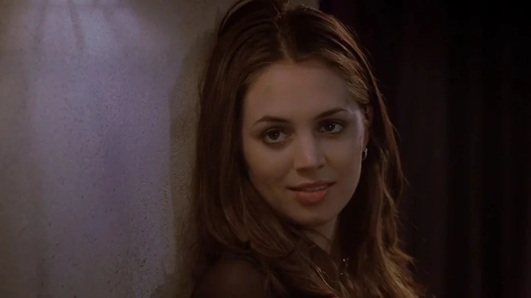 A young woman with long brown hair leans against a wall, looking slightly to the side with a subtle smile, in a dimly lit setting.