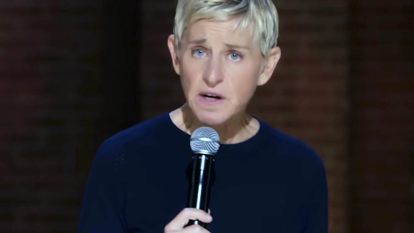 A person with short blonde hair, wearing a dark shirt, holds a microphone and appears to be speaking on stage with a blurred brick background.