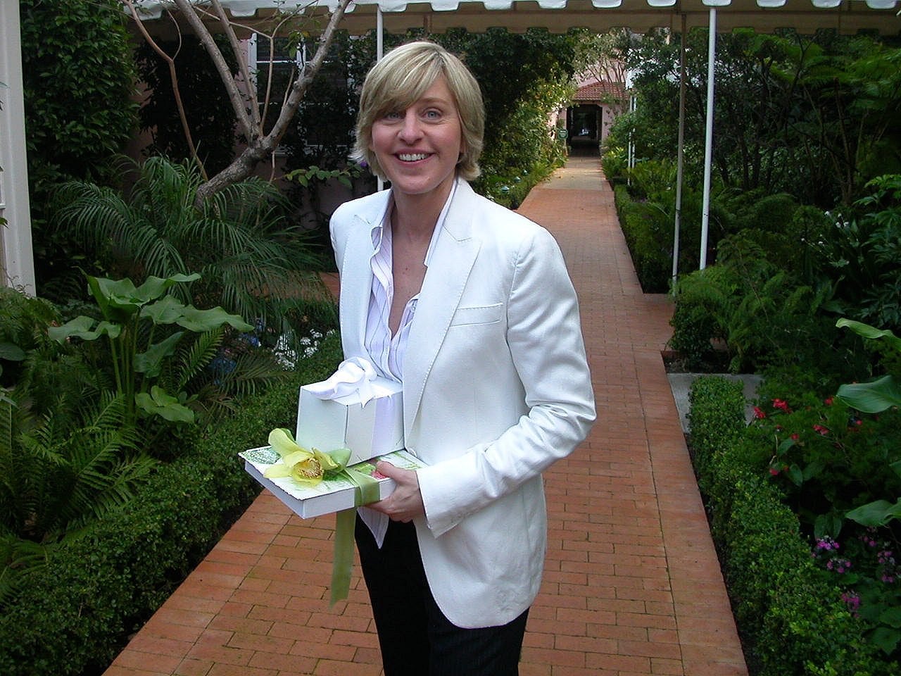 A person with short blonde hair, wearing a white blazer and light shirt, stands on a brick pathway in a lush garden, smiling and holding two gift boxes wrapped with ribbons.