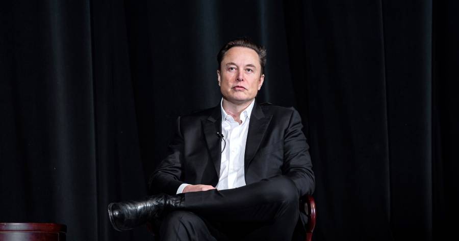 A man in a black suit and white shirt sits on a chair with legs crossed against a dark curtain background, looking forward with a neutral expression.