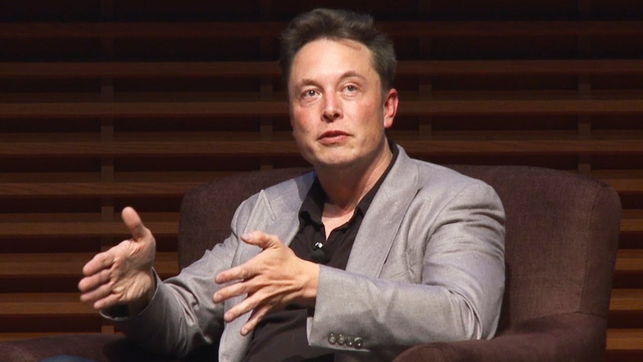A man in a gray blazer and black shirt sits in a brown armchair, gesturing with his hands while speaking. The background features horizontal wooden panels.
