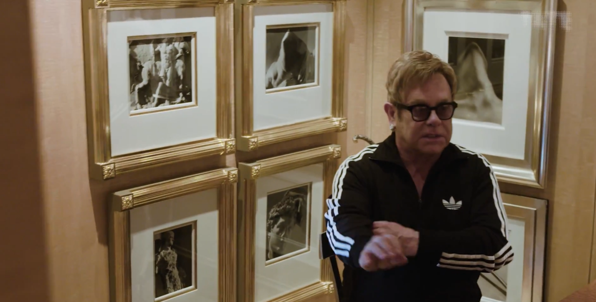 A man wearing glasses and an Adidas tracksuit sits in front of a wall decorated with several framed black-and-white photographs.