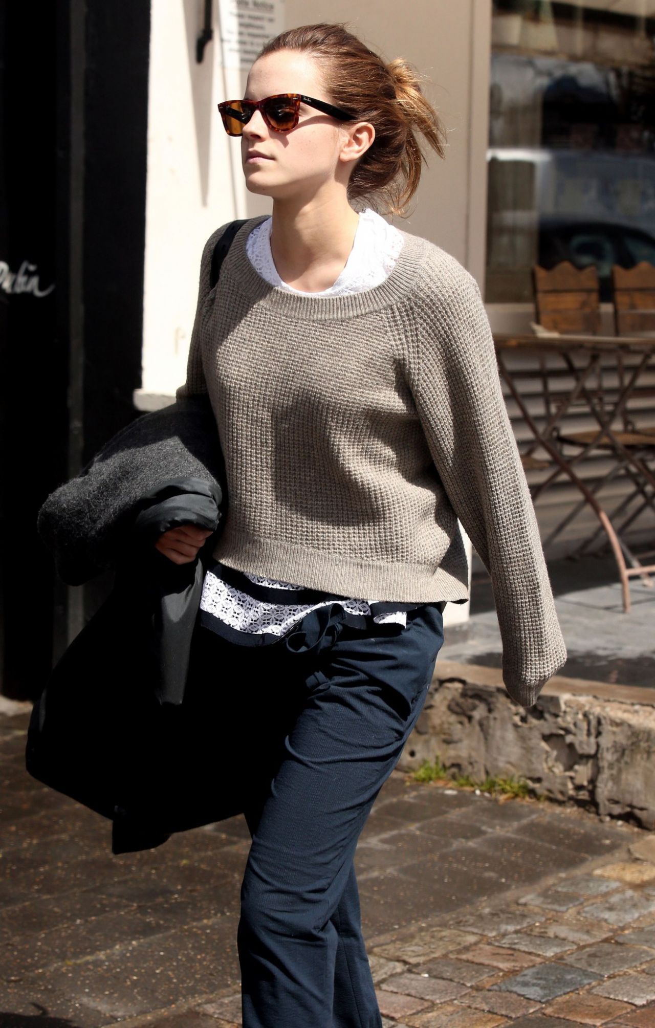 A woman wearing sunglasses, a beige sweater over a white top, and navy pants walks outside on a city street, holding a coat in one arm. There are empty chairs and tables in the background.