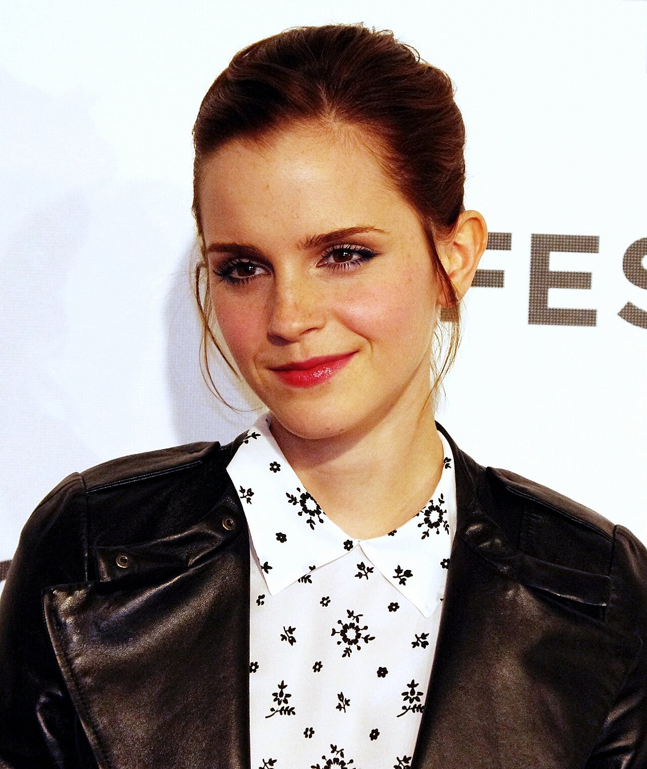 A woman with fair skin and brown hair tied back, wearing a black leather jacket over a white collared shirt with a black floral pattern, stands in front of a white background with partial text. She is smiling softly.