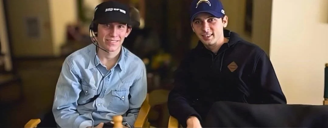Two young men sitting indoors on chairs, smiling at the camera. One wears a headset and a light blue shirt, while the other wears a dark jacket and a blue cap with a sports team logo. The background is softly blurred.