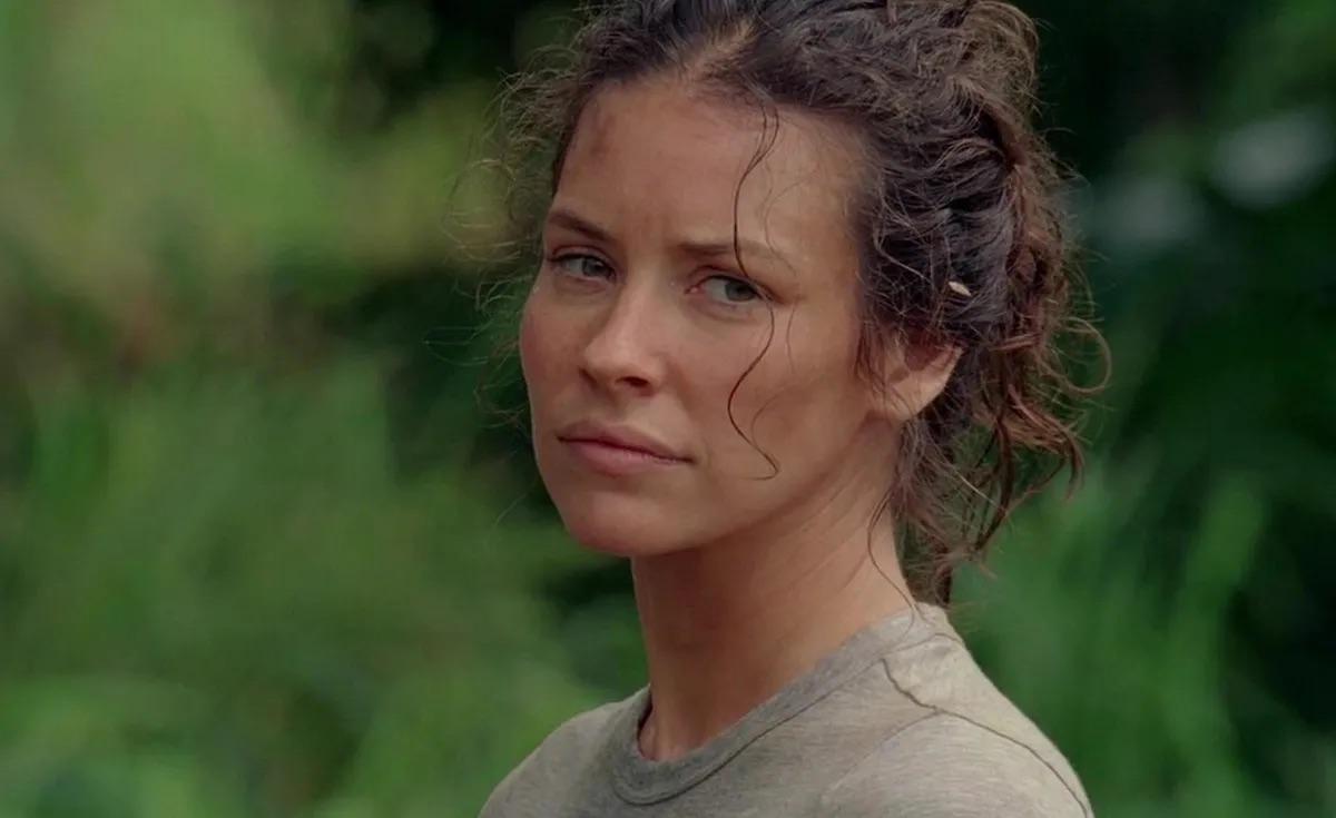 A woman with curly brown hair, wearing a light gray t-shirt, stands outdoors with a serious expression. Lush green foliage is blurred in the background.