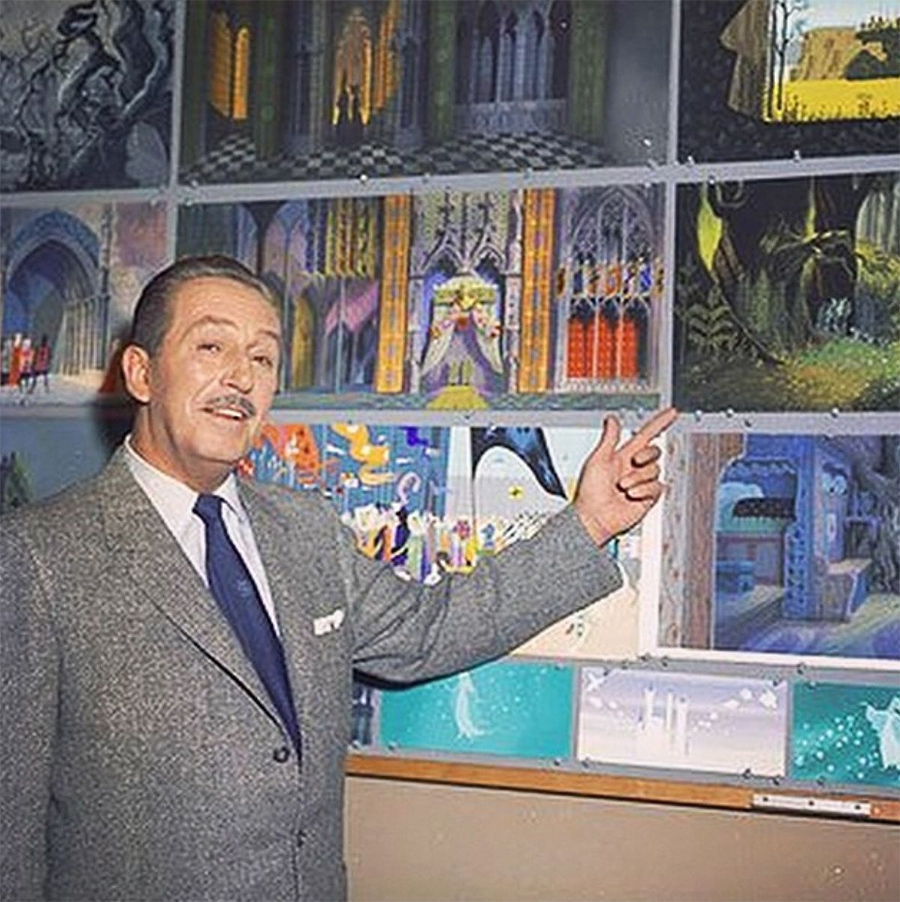 A man in a suit with a mustache stands in front of a wall covered in colorful concept art, smiling and gesturing toward the artwork with his right hand.