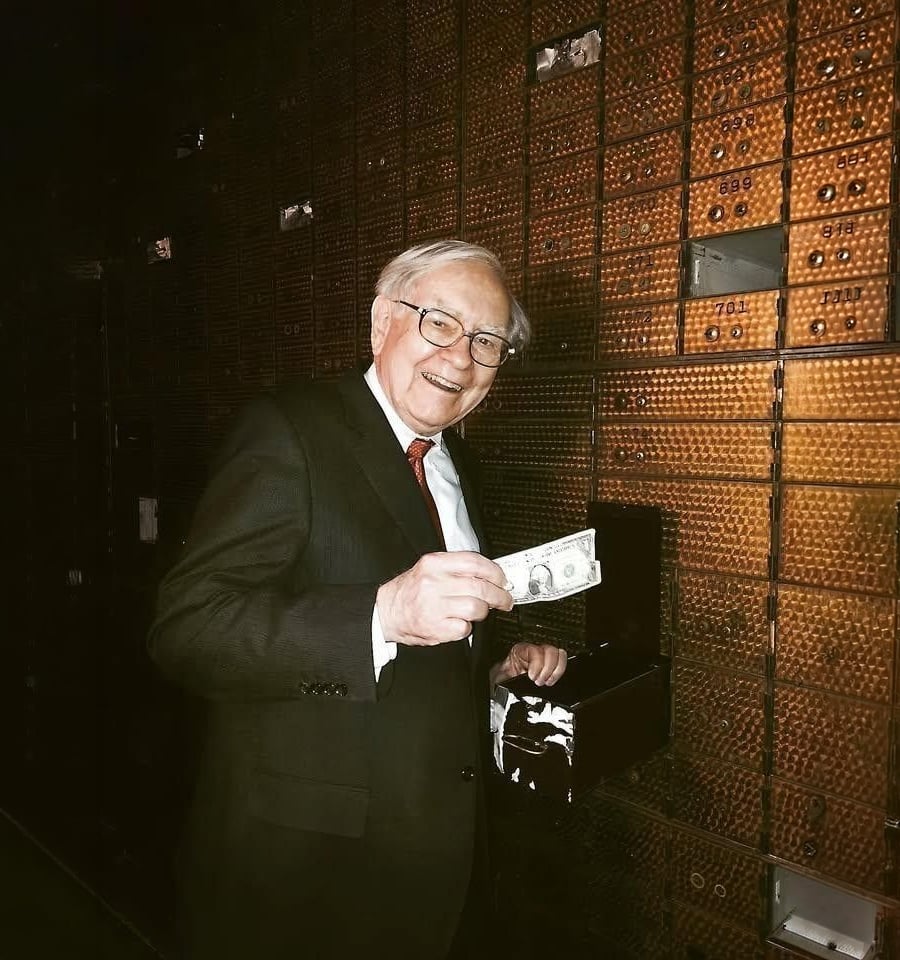 An elderly man in a suit and glasses smiles while holding a U.S. dollar bill in front of an open safe deposit box inside a bank vault with metallic safety deposit boxes behind him.