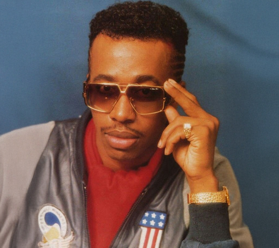 A man wearing gold sunglasses, a gold watch, and a grey jacket with American flag and patch details poses against a blue background, touching his glasses and looking confidently at the camera.
