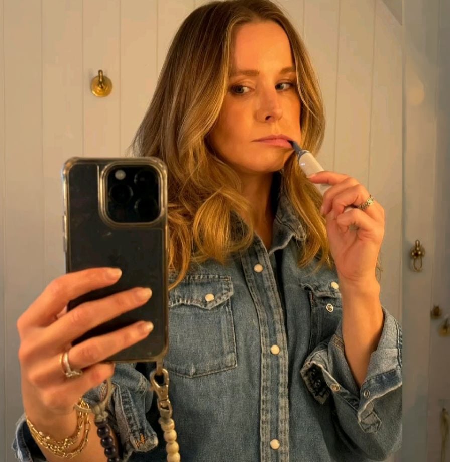 A woman wearing a denim shirt takes a mirror selfie while holding a lipstick to her lips, with her smartphone visible in one hand. Warm indoor lighting creates a cozy atmosphere.
