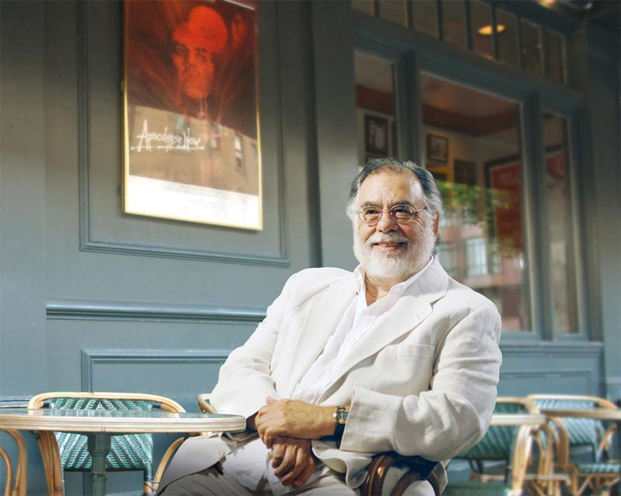 An older man with white hair and a beard, wearing glasses and a white suit, sits smiling at an outdoor cafe table. Behind him, a movie poster for "Apocalypse Now" is visible on a blue-gray wall.