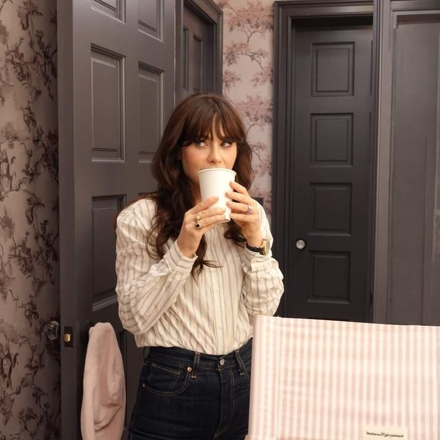 A woman with long brown hair, wearing a striped shirt and jeans, stands in a bathroom with patterned wallpaper and dark doors, sipping from a white cup. A pink and white striped object is in the foreground.