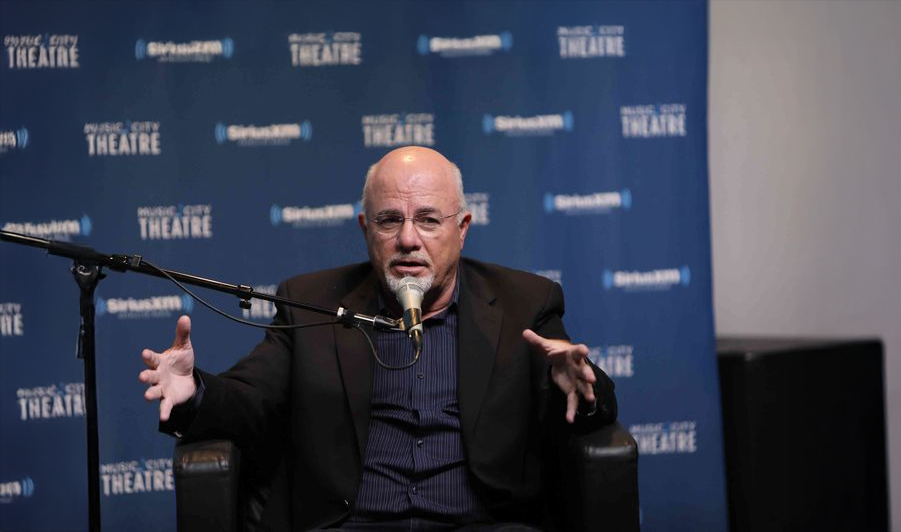A bald man with glasses and a goatee speaks into a microphone while gesturing with his hands, seated in front of a blue backdrop with "Music City Theatre" and "SiriusXM" logos.