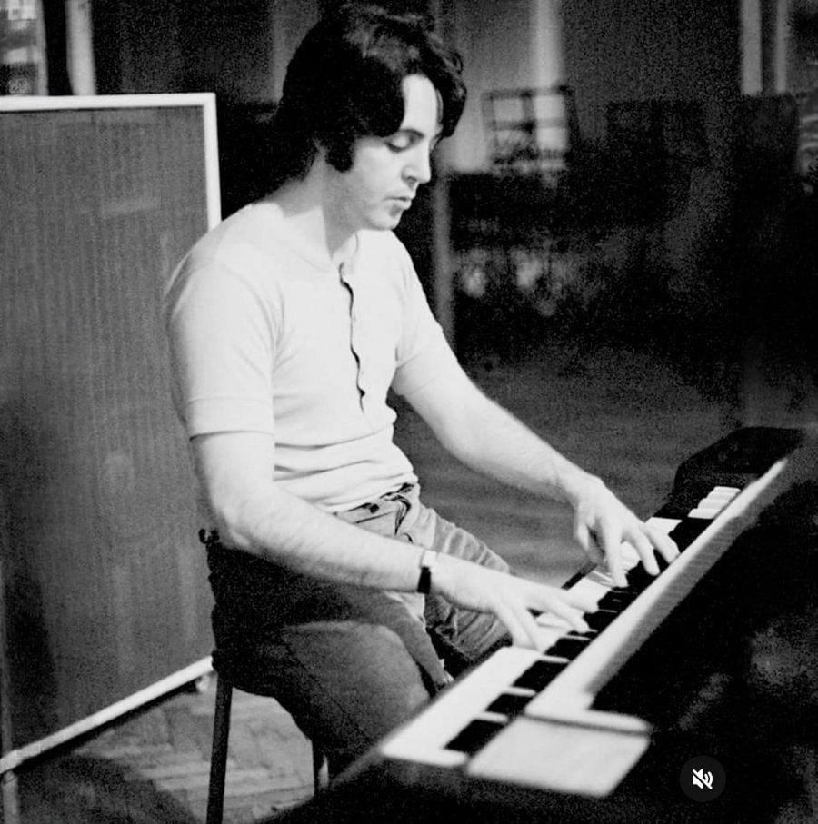 A man with dark hair sits on a stool playing an electric piano in a dimly lit room, focused on the keys. The scene is in black and white, with a blurred background and music equipment nearby.