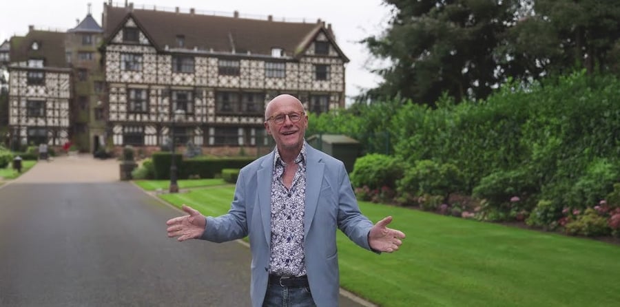 A smiling man in a light blue blazer and patterned shirt stands with open arms on a paved driveway, with a large historic timber-framed building and manicured lawns in the background.