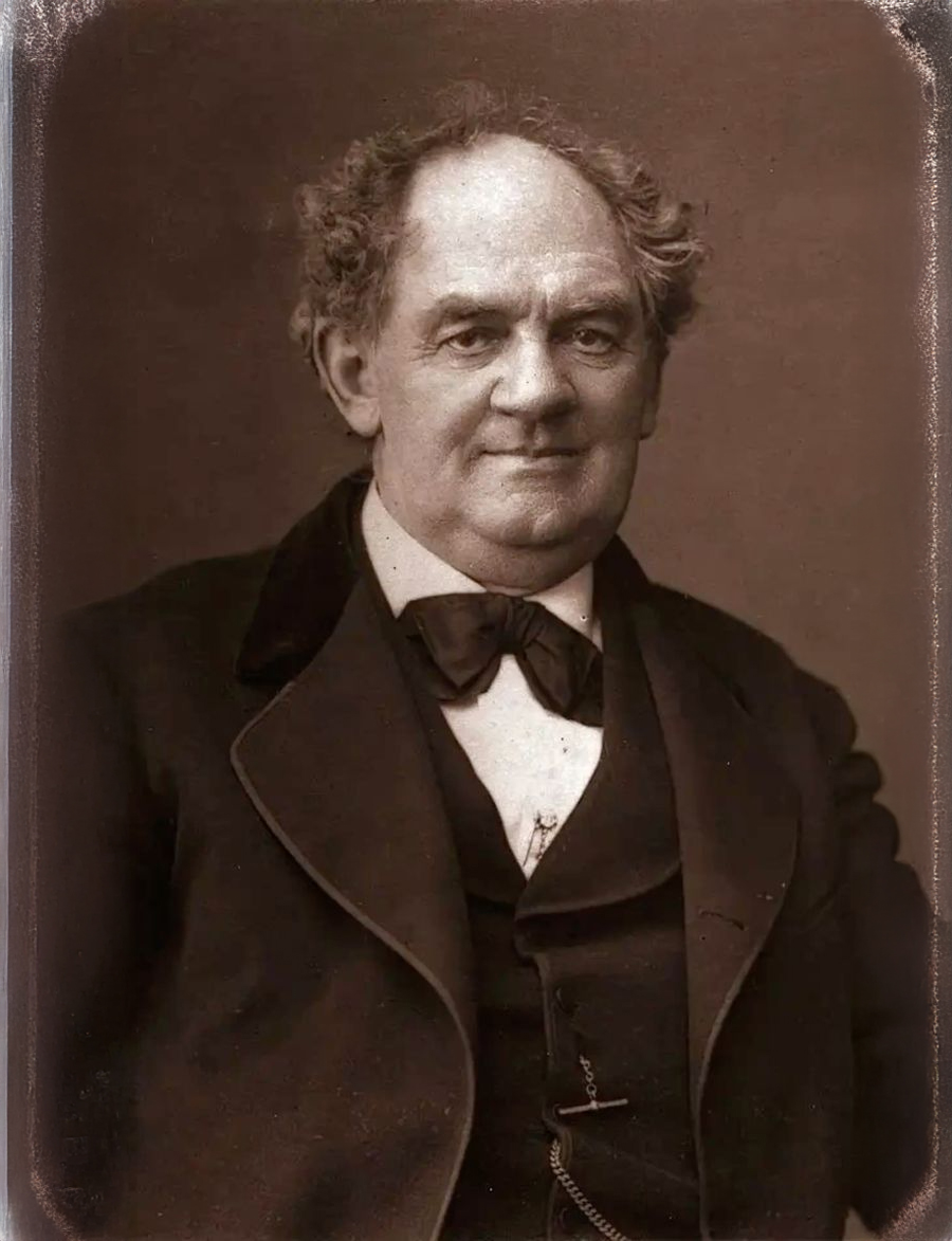 A sepia-toned portrait of an older man with curly hair, wearing a dark suit, white shirt, and bow tie, looking directly at the camera with a slight smile.