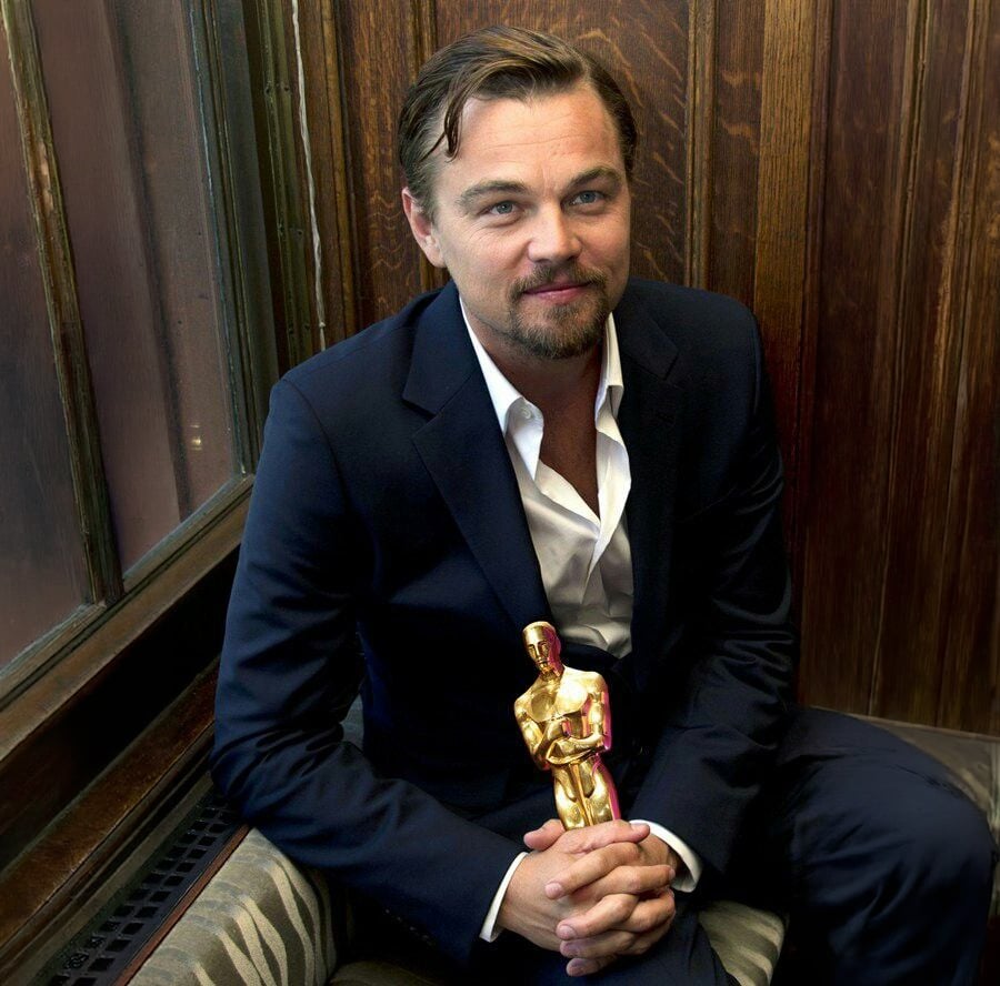 A man in a dark suit, with a beard and light brown hair, sits on a cushioned bench holding a gold Oscar statuette, smiling slightly, against a wood-paneled wall.