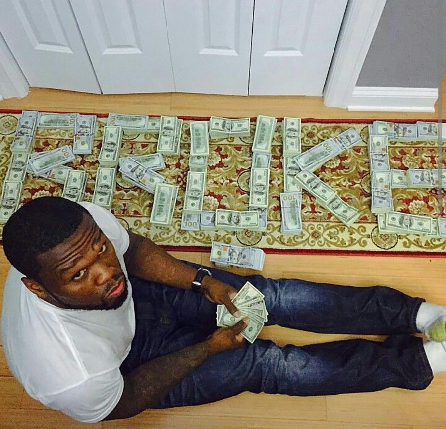 A man in a white t-shirt sits on the floor holding cash, with stacks of dollar bills arranged on a rug to spell the word "BROKE.