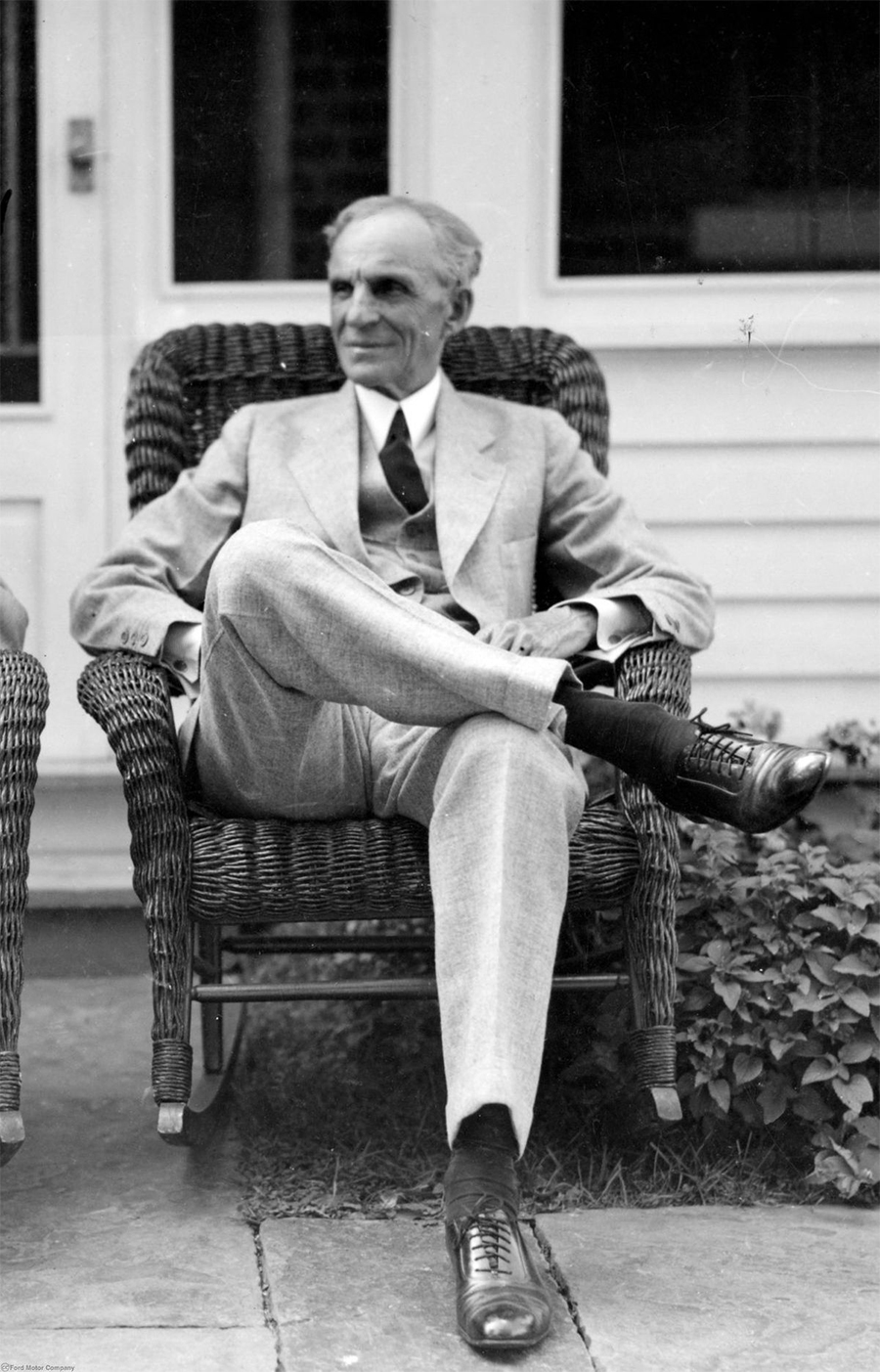 An older man in a suit sits with legs crossed in a wicker chair on a porch, looking to the side. The setting appears relaxed, with plants growing beside the chair and a white building in the background.
