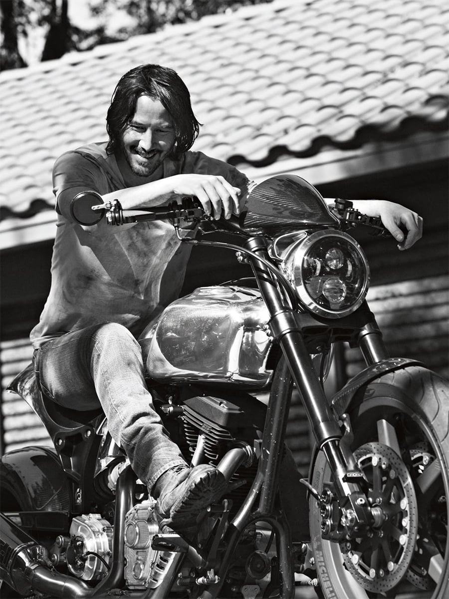 A man with long hair and a beard smiles while sitting on a motorcycle. He leans forward, holding the handlebars, and wears a casual t-shirt and jeans. The background features a tiled roof and horizontal wooden panels.
