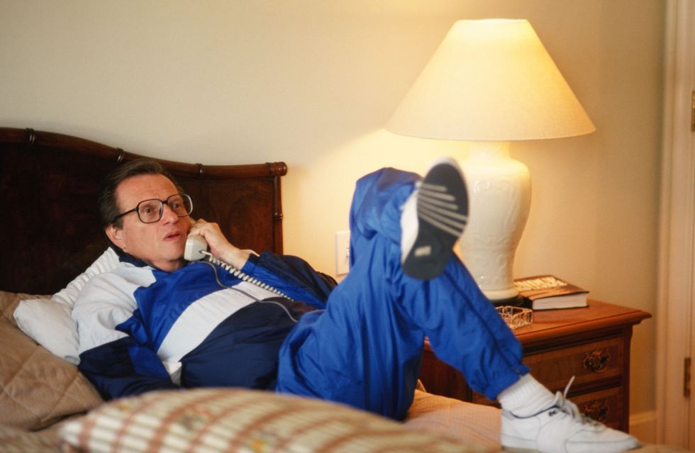 A person in a blue and white tracksuit reclines on a bed, holding a corded phone to their ear. One leg is propped up, and a lamp and books are visible on the bedside table nearby.