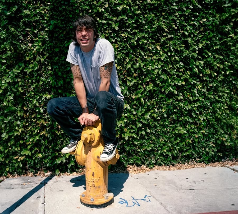 A man in a gray t-shirt and black jeans squats on top of a yellow fire hydrant, with a leafy green hedge behind him and a sidewalk beneath. He is smiling and wearing black sneakers.