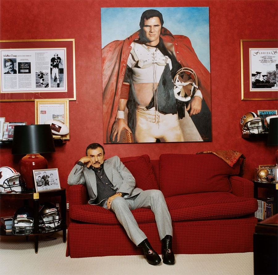 A man in a gray suit sits on a red couch in a room with red walls, below a large portrait of himself in a football uniform with a cape. The room is decorated with sports memorabilia and framed photos.