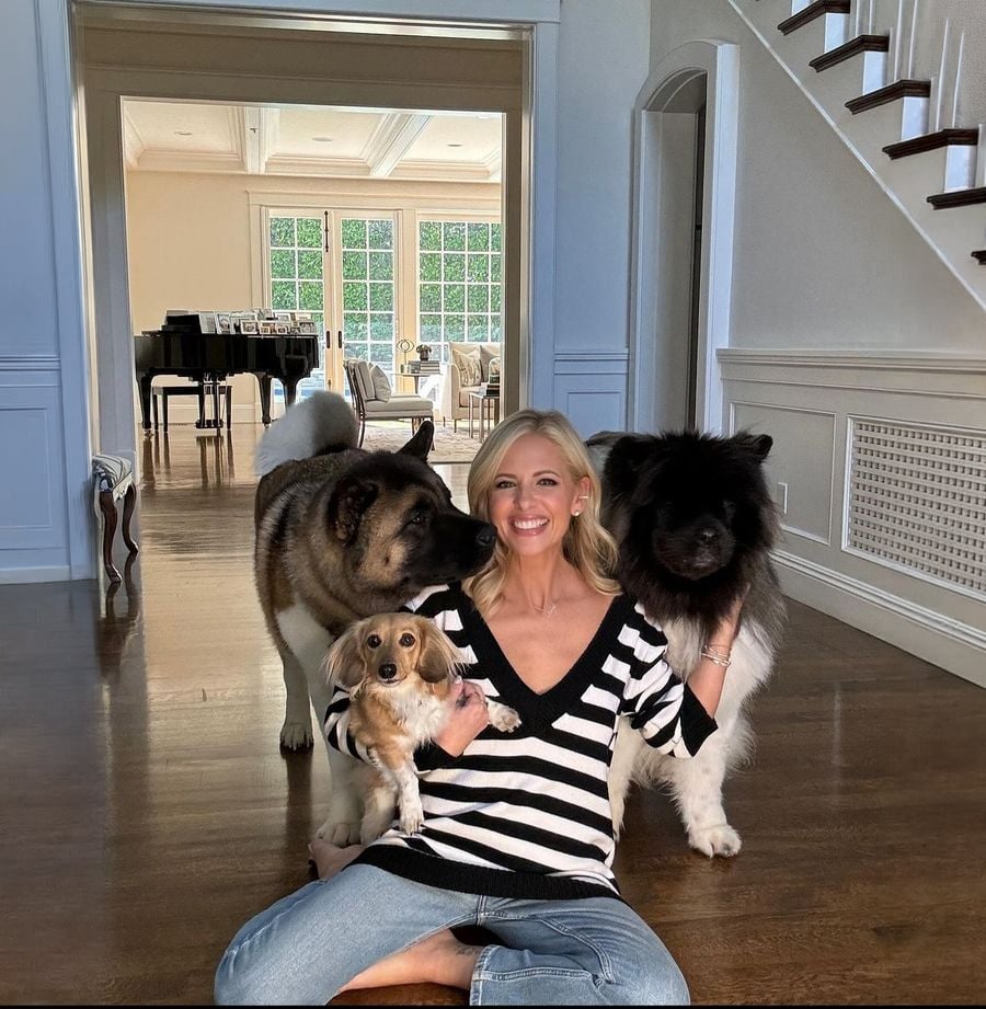 A smiling woman sits on a wooden floor, holding a small dog in her lap, with two large fluffy dogs beside her. Behind them is a bright room with a piano, chairs, and large windows.