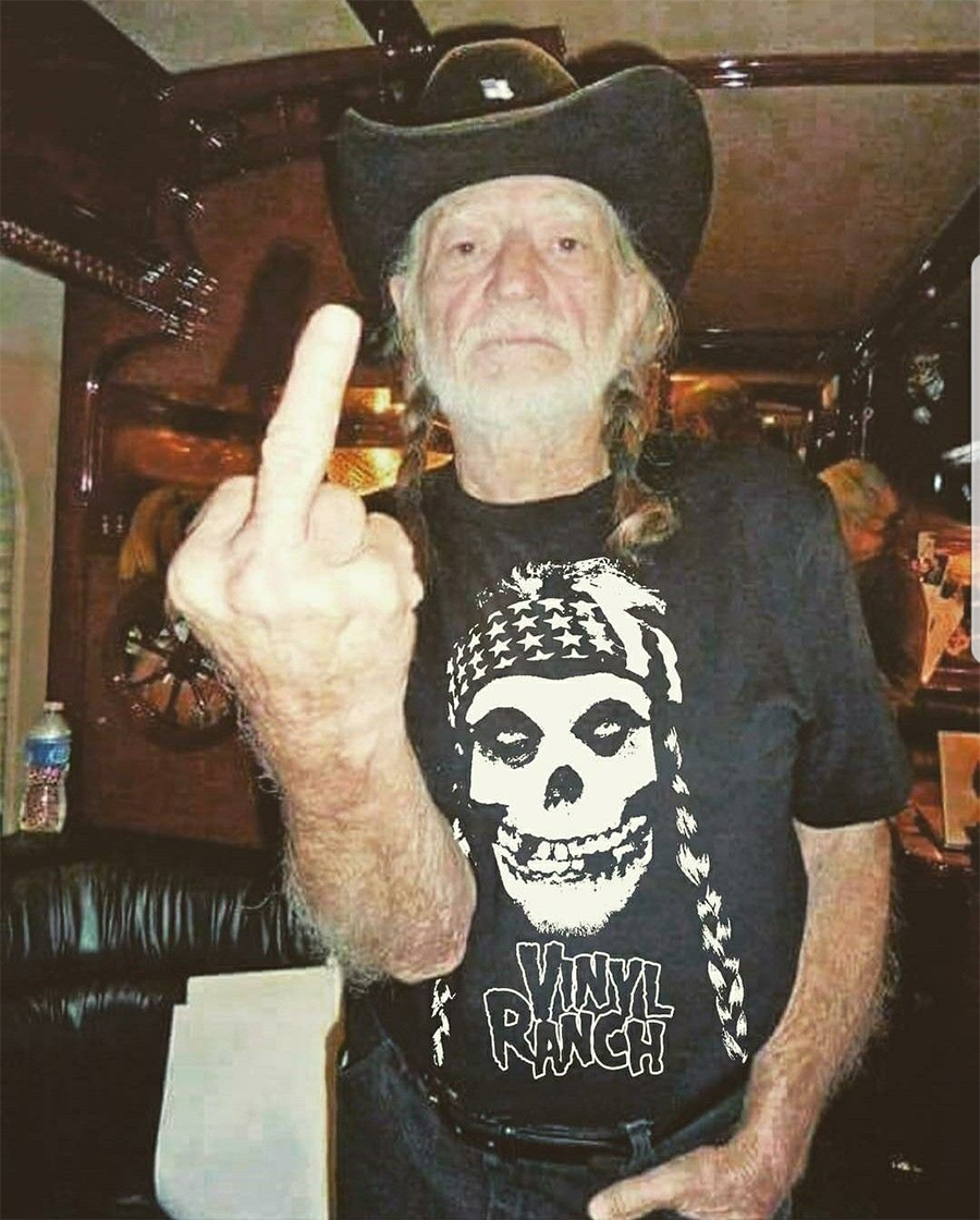 An older man with long hair in braids, wearing a black cowboy hat and a black "Vinyl Ranch" t-shirt, stands indoors and raises his middle finger toward the camera.