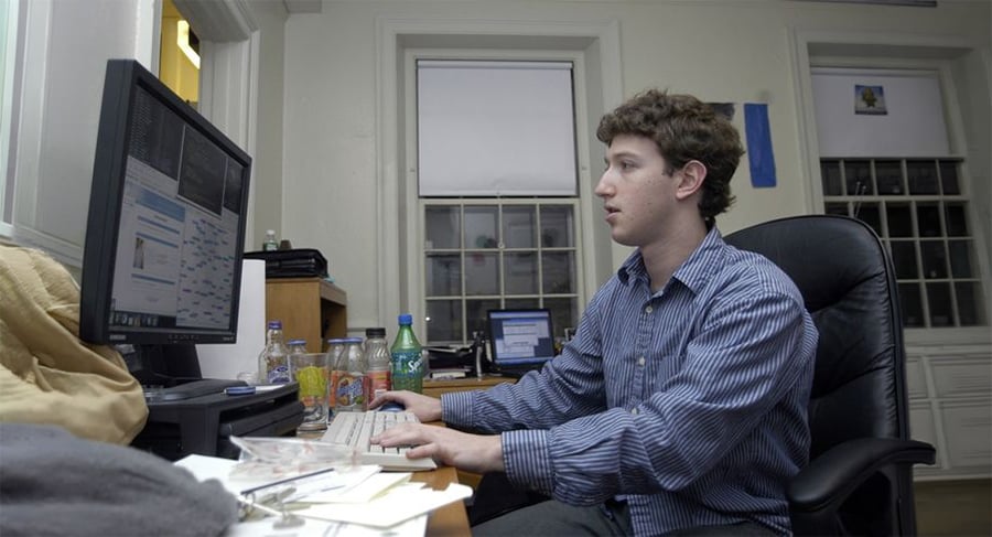 A young man in a blue striped shirt sits at a desk, typing on a computer keyboard. The desk is cluttered with papers and bottles, and there are two monitors and a laptop in a room with large windows.