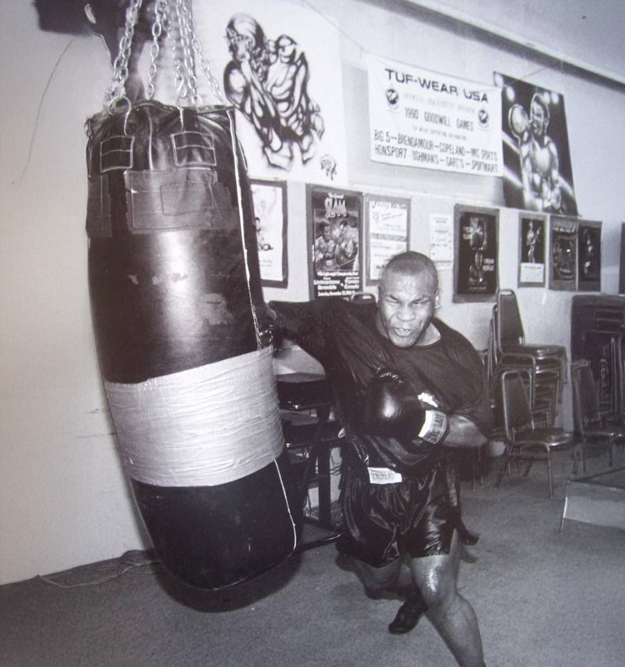 A boxer in gloves and workout attire powerfully punches a heavy bag in a gym filled with posters and stacked chairs. The action is dynamic, showing strength and focus. The image is in black and white.