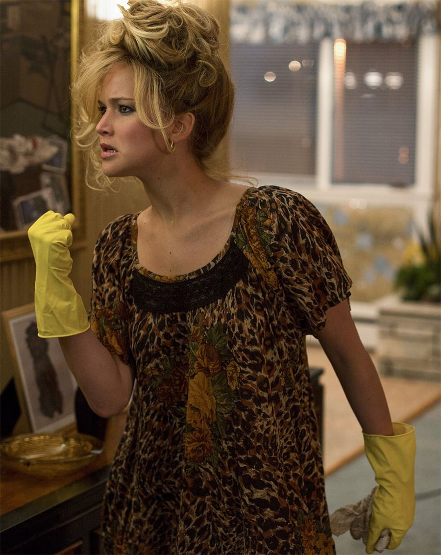 A woman wearing yellow rubber cleaning gloves and a leopard-print dress stands indoors, looking tense and determined. Her blonde hair is styled in a messy updo, and she appears to be in the middle of cleaning.