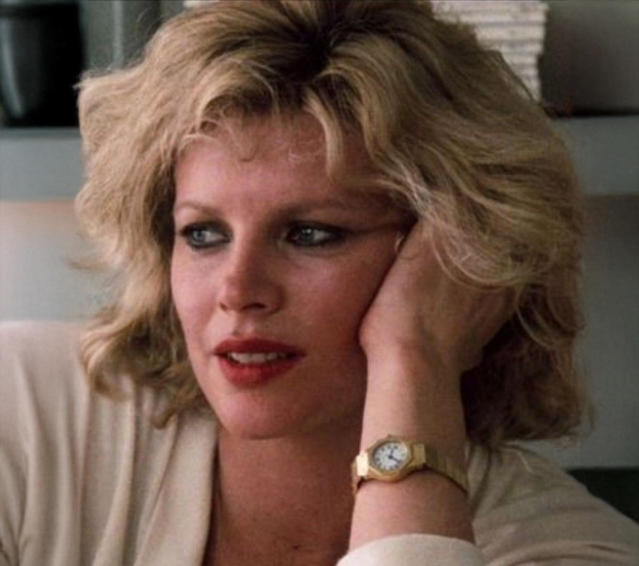 A woman with blonde, wavy hair rests her head on her hand, looking thoughtfully to the side. She wears a light-colored top, dark eyeliner, and a gold wristwatch. The background is softly blurred.
